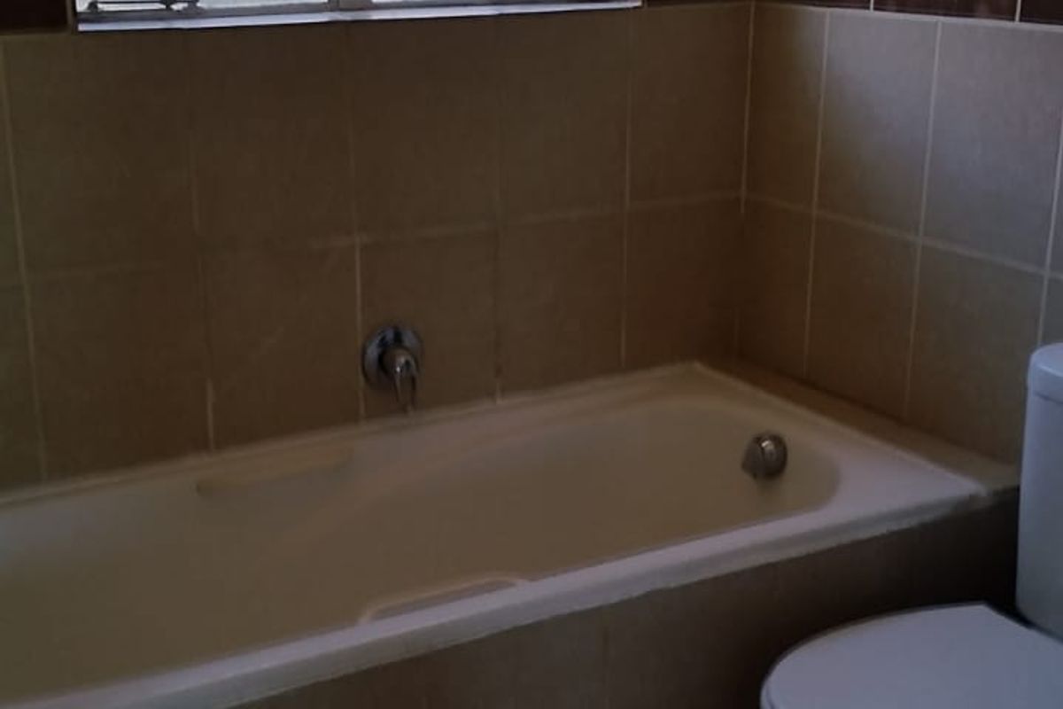 A functional bathroom featuring a built-in bathtub, toilet, and neutral wall and floor tiling with security bars on the window.