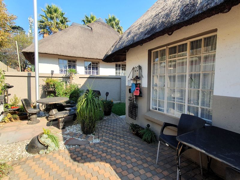 A rear exterior view featuring a paved patio area, thatched roof architecture, and a surrounding boundary wall.