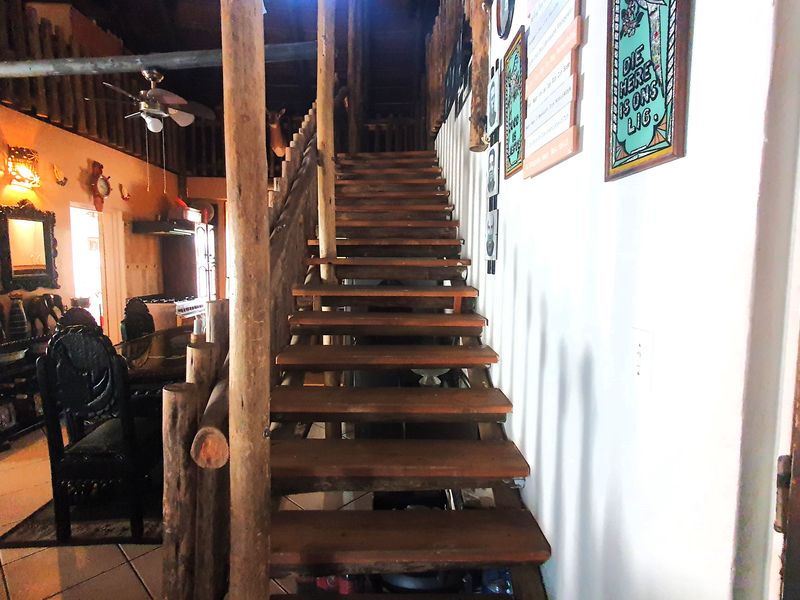 A rustic interior hallway featuring a prominent timber staircase with log support posts and tiled flooring, leading to an open-plan living area.