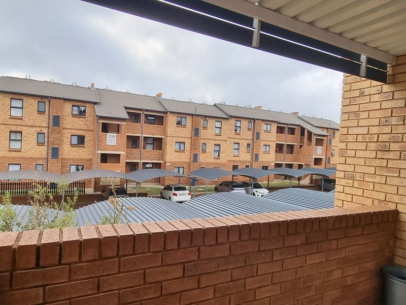 A covered balcony featuring a brick balustrade and metal roofing, overlooking the complex parking area and neighboring townhouse blocks.
