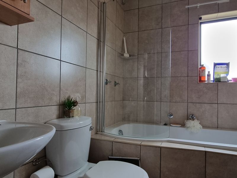 A functional bathroom featuring a pedestal basin, toilet, and a combined bathtub with a glass shower screen, finished with neutral wall and floor tiles.