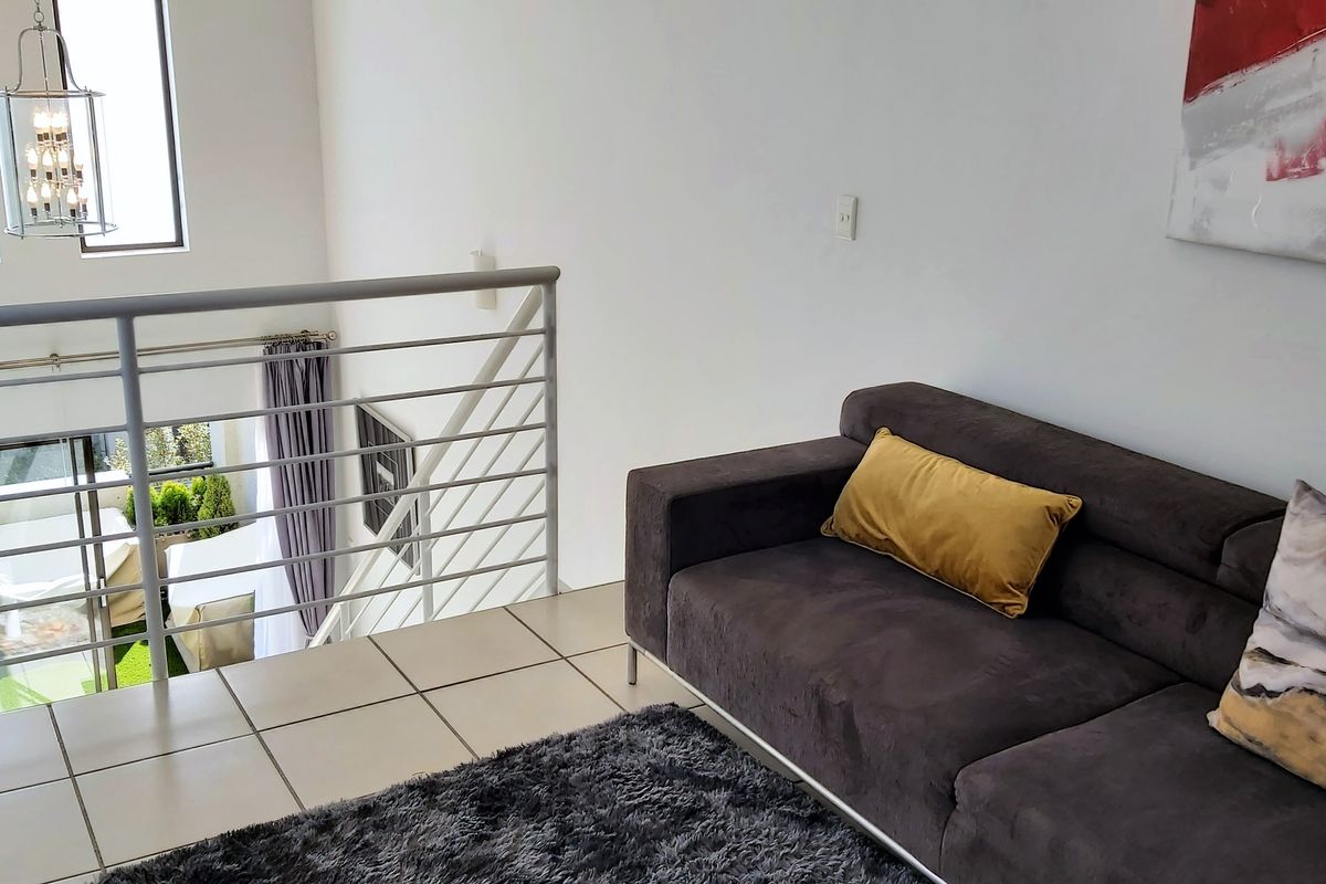 A mezzanine living area featuring tiled flooring, a modern metal balustrade, and a double-volume ceiling with a stylish pendant light fixture.