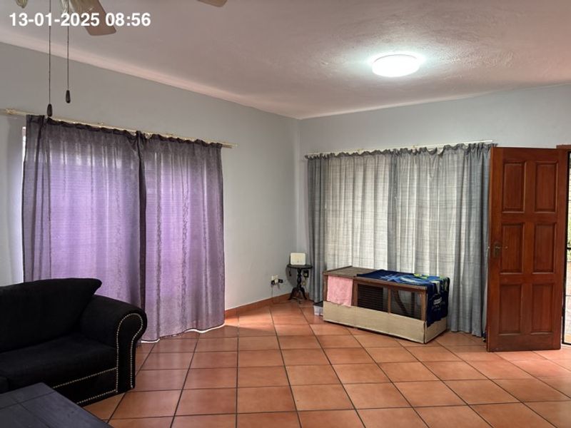 A living room featuring terracotta floor tiles, a ceiling fan, and natural light through large windows with curtains.