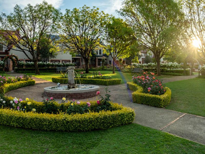 A beautifully landscaped communal garden featuring a central stone fountain, manicured hedges, paved walkways, and mature trees in a peaceful residential setting.