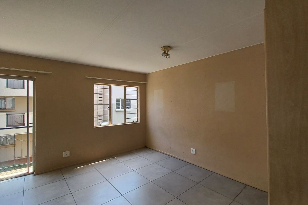 An empty living room featuring neutral tiled flooring, a sliding glass door with access to a balcony, and security bars on the window.