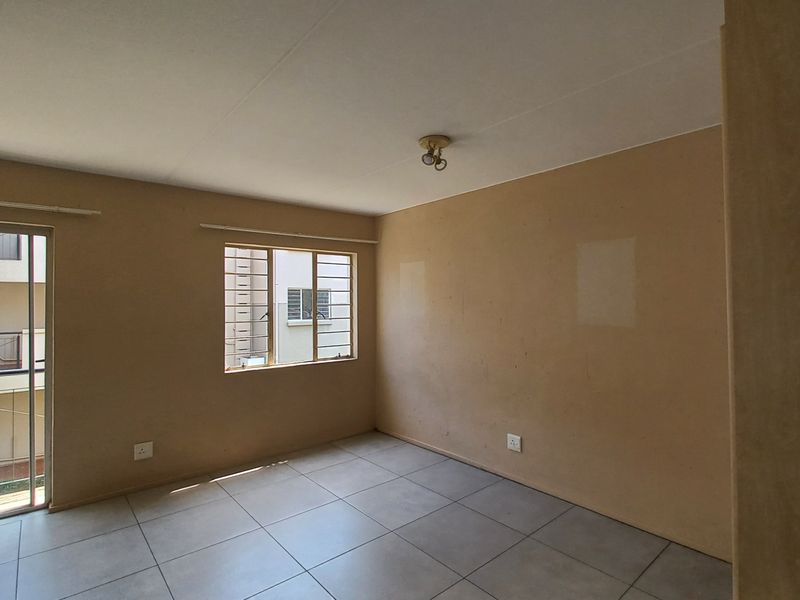 An empty living room featuring neutral tiled flooring, a sliding glass door with access to a balcony, and security bars on the window.
