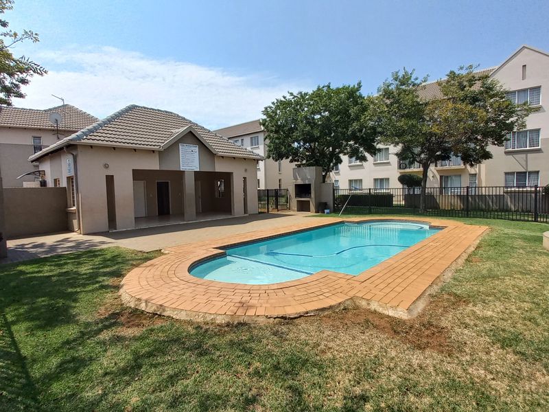 Communal pool area featuring a swimming pool with brick coping, adjacent clubhouse with built-in braai, and landscaped garden surroundings within a secure complex.