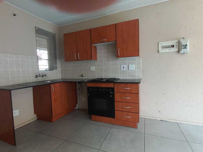 A compact kitchen featuring wooden cabinetry, a built-in oven and hob, tiled backsplash, and tiled flooring. The space is currently vacant.