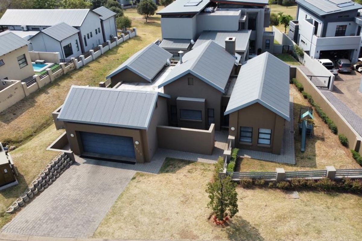 Aerial view of a modern residential property featuring a metal roof, paved driveway, attached garage, and landscaped garden area.