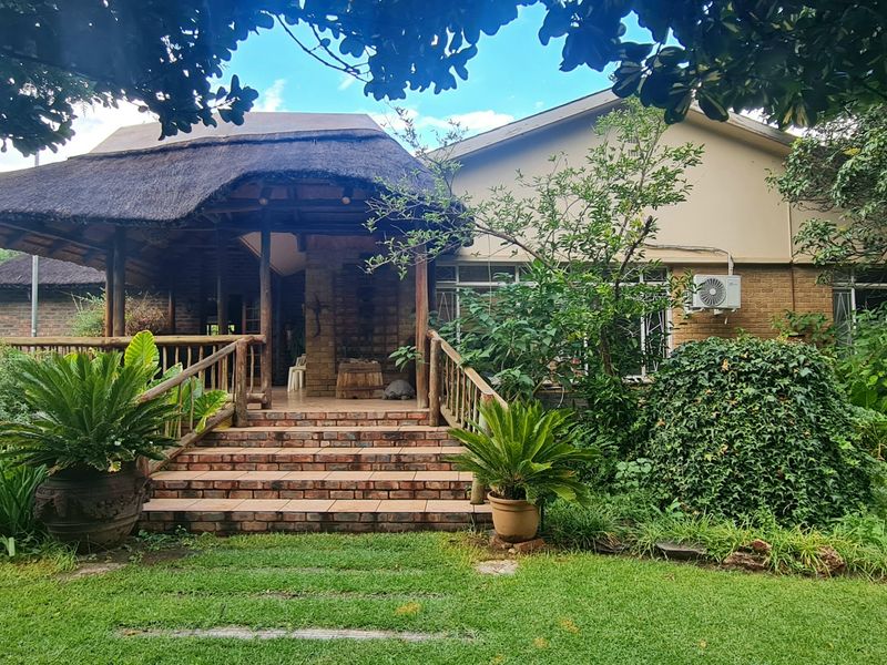 A rear view of the house featuring a covered patio with a traditional thatch roof, brick staircase, and established garden surroundings.