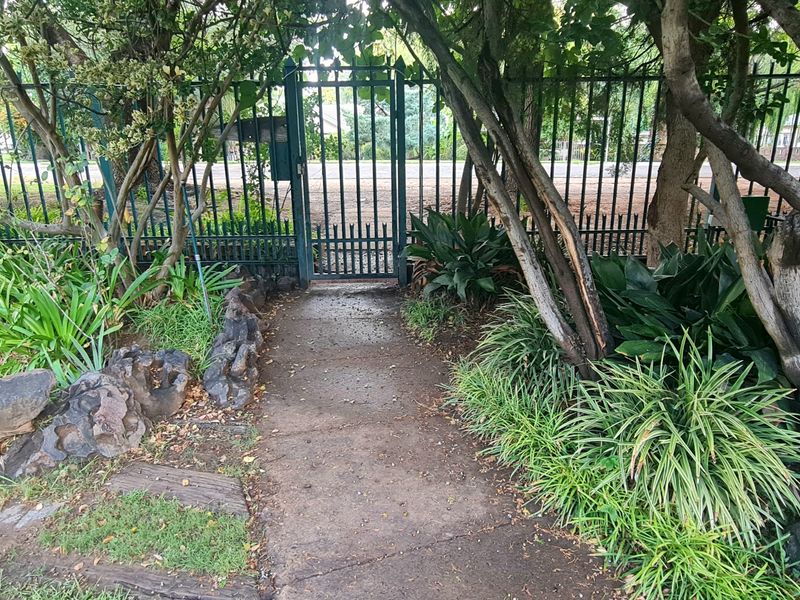 A front garden entrance featuring a concrete pathway leading to a metal security gate and perimeter fencing, surrounded by established trees and garden beds.