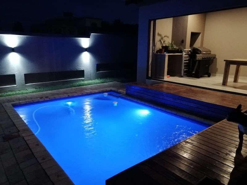 Night view of a swimming pool with timber decking, paved surrounds, and integrated wall lighting, adjacent to a covered patio area.