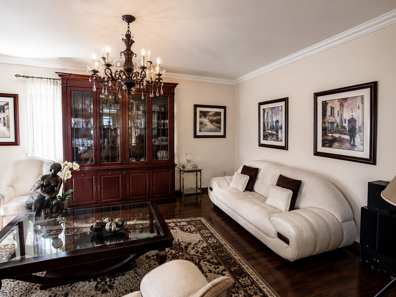 A well-maintained living room featuring rich hardwood flooring, elegant crown molding, a classic chandelier, and a large built-in wooden display cabinet.