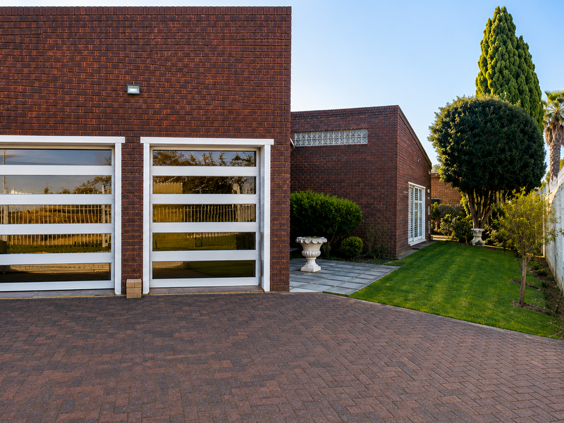 Modern brick-faced home exterior featuring a double garage with glass panel doors, a paved driveway, and a well-maintained garden with a patio area.