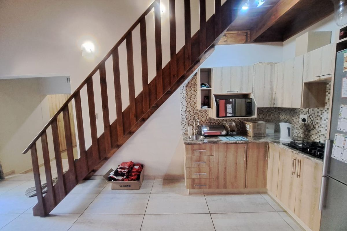 Compact kitchen area featuring light wood cabinetry, mosaic backsplash, and gas hob, situated beneath a wooden staircase with tiled flooring throughout.