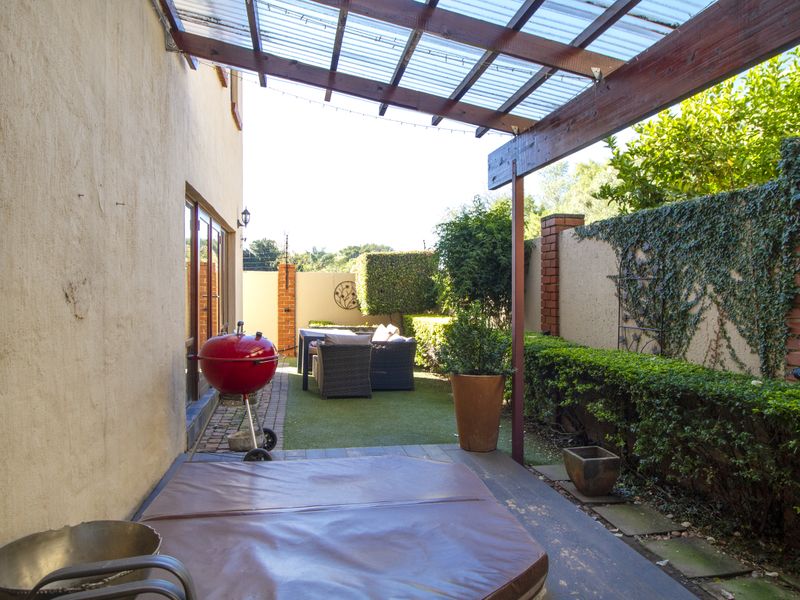 A covered outdoor patio area featuring a timber pergola with polycarbonate roofing, overlooking a landscaped garden with brick boundary walls and artificial lawn.
