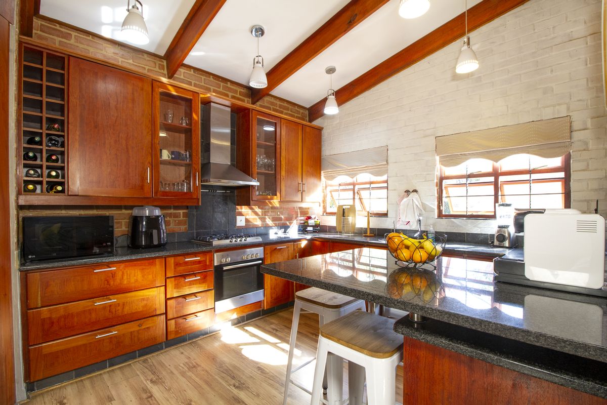 A warm, rustic-style kitchen featuring rich wooden cabinetry, granite countertops, exposed ceiling beams, and a brick feature wall with integrated appliances.