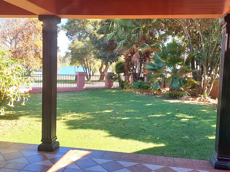 A covered patio featuring decorative columns and tiled flooring, overlooking a private garden with established trees and perimeter fencing.