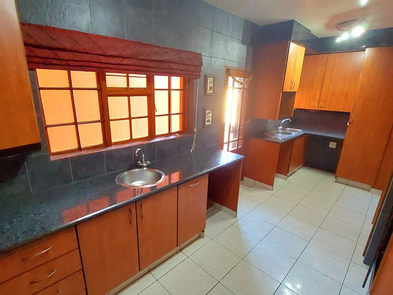 A functional kitchen featuring wooden cabinetry, dark tiled splashbacks, double stainless steel sinks, and light-colored floor tiles in a clean, vacant space.