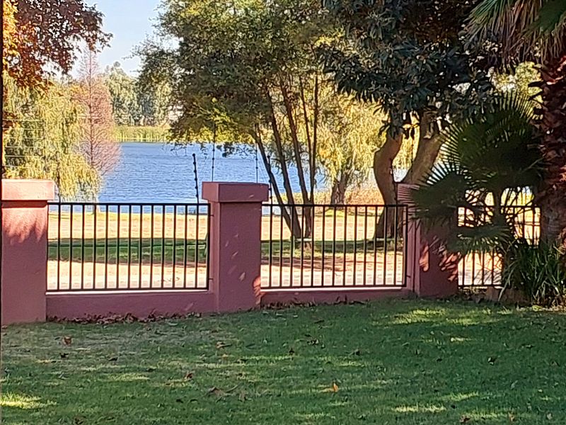 A scenic view of a lake framed by mature trees, featuring a well-maintained garden with masonry pillars and metal perimeter fencing.