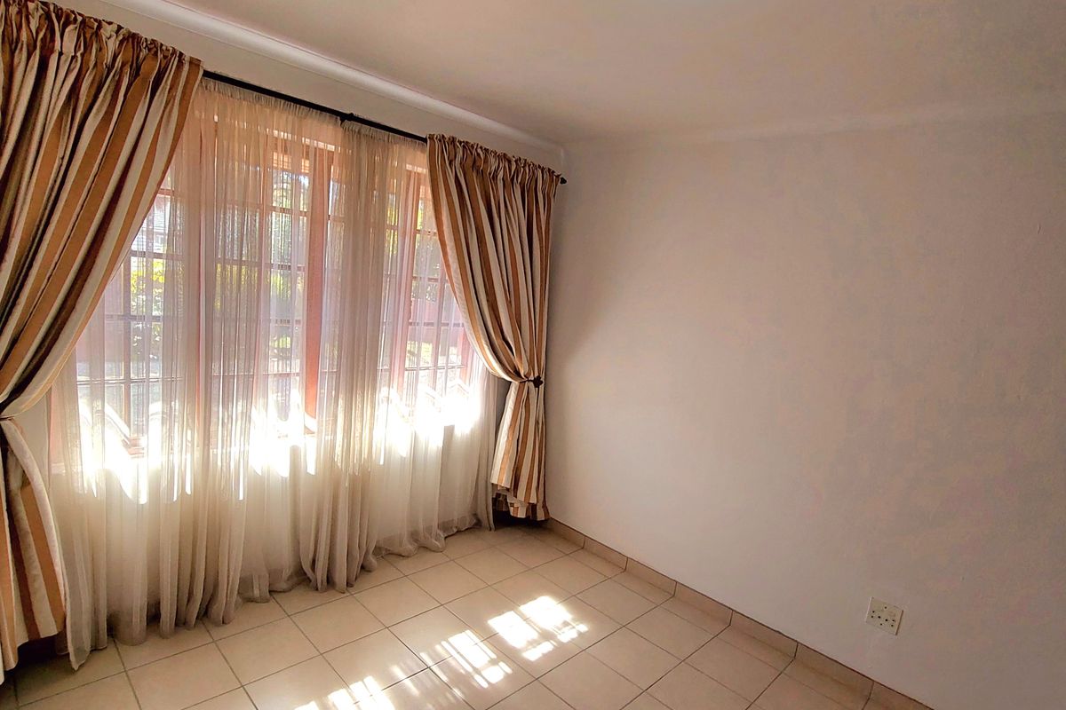A bright, empty bedroom featuring neutral tiled flooring, a ceiling light fixture, and large windows with curtains.