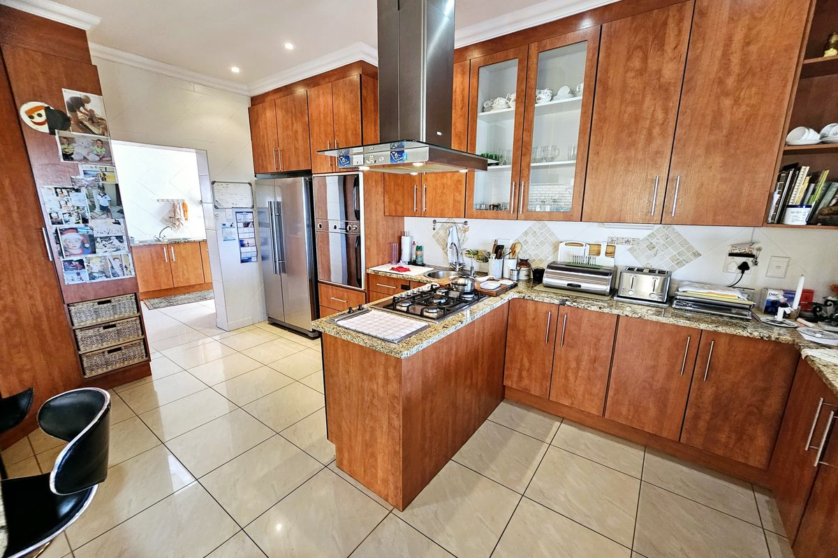 Spacious kitchen featuring warm-toned timber cabinetry, granite countertops, a central gas hob with extractor, and tiled flooring, with an adjacent scullery area.