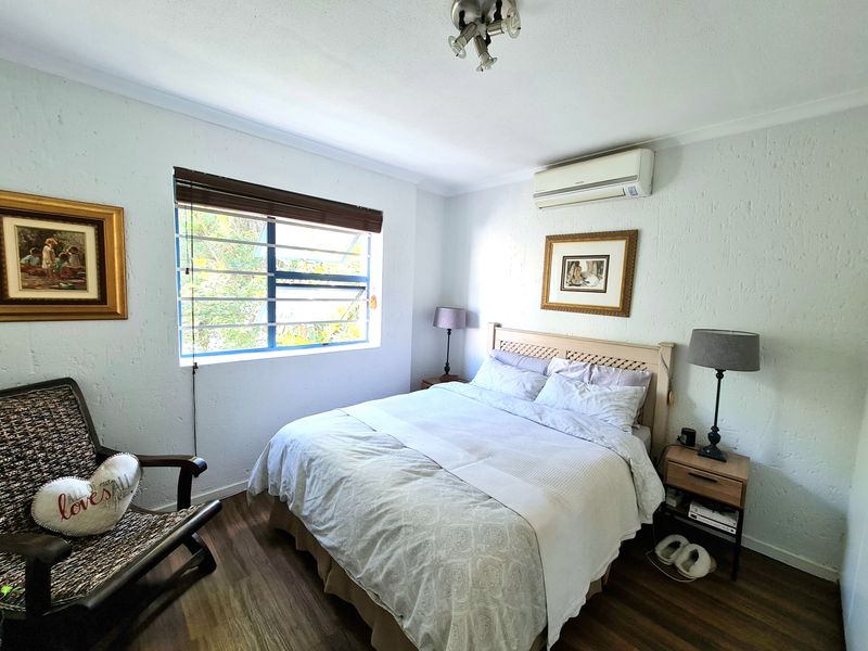 A cozy bedroom featuring laminate flooring, a split-system air conditioner, and a louvered window with security bars.