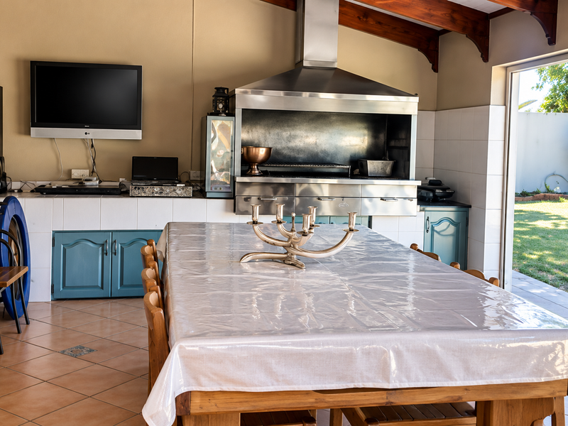Covered outdoor patio featuring a built-in braai with stainless steel extractor, tiled flooring, and exposed timber beams, opening directly onto the garden.