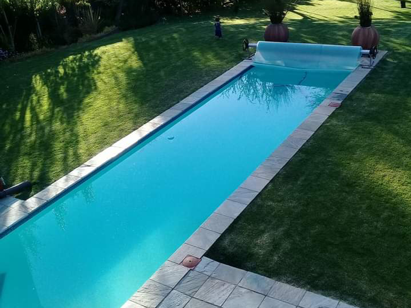 A long, narrow swimming pool set within a well-maintained garden, featuring stone paving and a retractable pool cover.