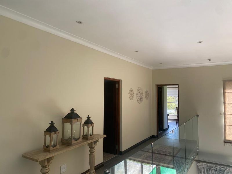 A bright and well-maintained hallway featuring a modern glass balustrade, recessed downlighting, and neutral wall finishes, providing access to adjacent rooms.