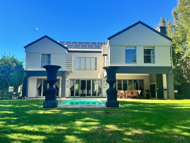 Rear view of a two-story house featuring a swimming pool, covered patio with built-in braai, and roof-mounted solar panels set in a landscaped garden.