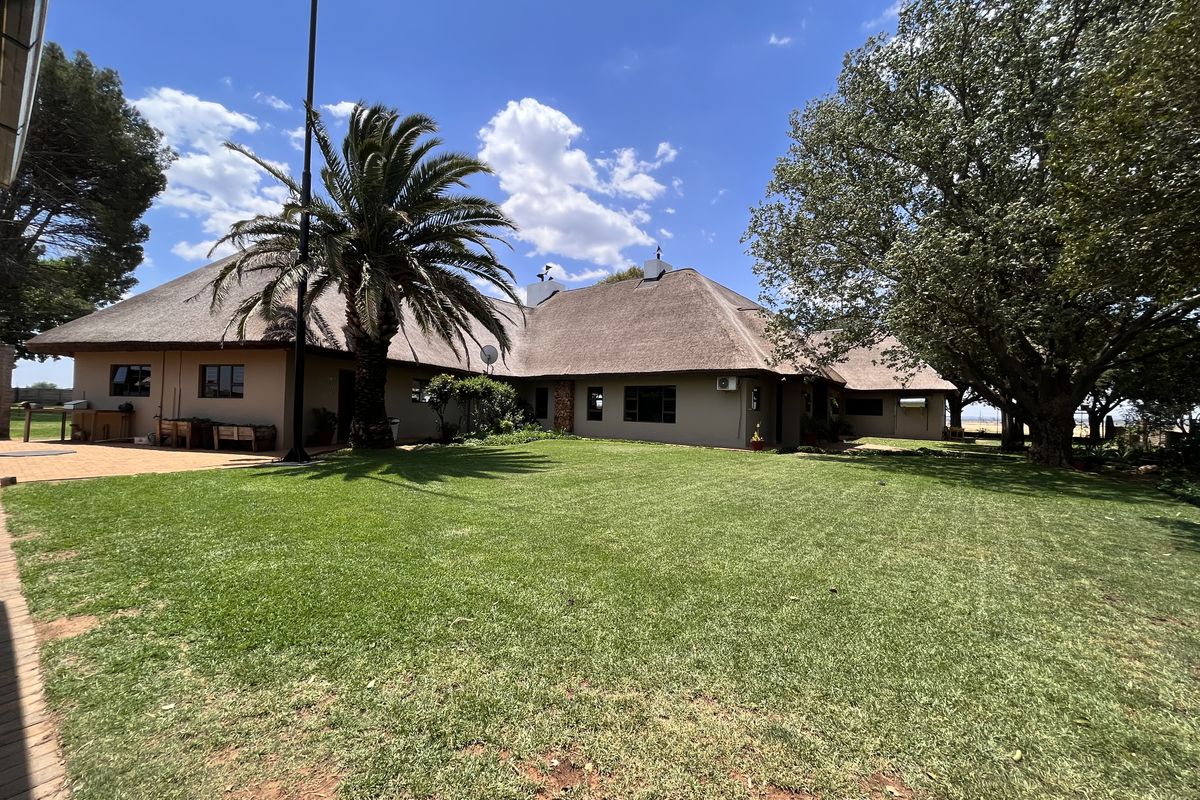 A spacious rear garden view featuring a well-maintained lawn, mature trees, and a large thatched-roof dwelling with a paved patio area.