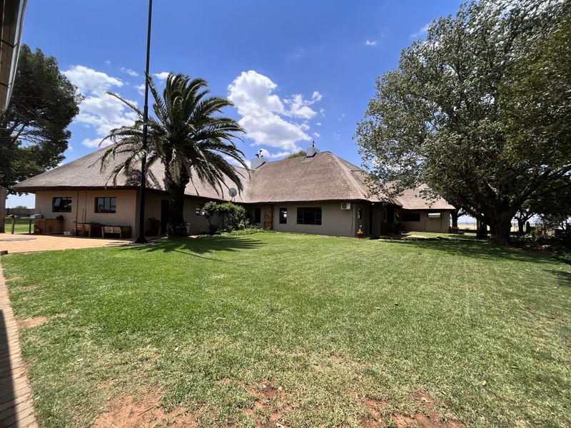 A spacious rear garden view featuring a well-maintained lawn, mature trees, and a large thatched-roof dwelling with a paved patio area.