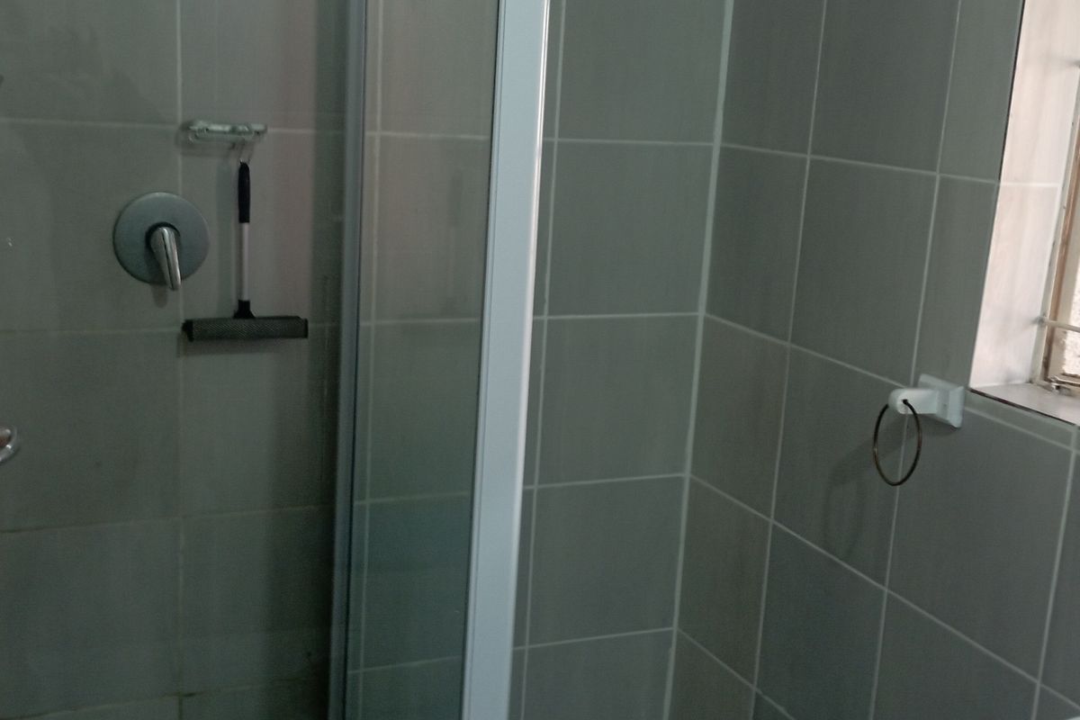 A functional bathroom shower area featuring grey wall tiles, a glass shower enclosure, and a window with security bars.