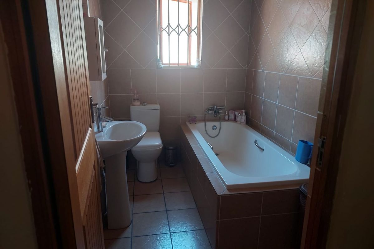 A functional bathroom featuring a pedestal basin, toilet, and a built-in bathtub with tiled finishes and a window with security bars.