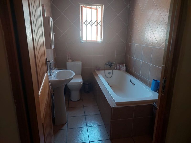 A functional bathroom featuring a pedestal basin, toilet, and a built-in bathtub with tiled finishes and a window with security bars.