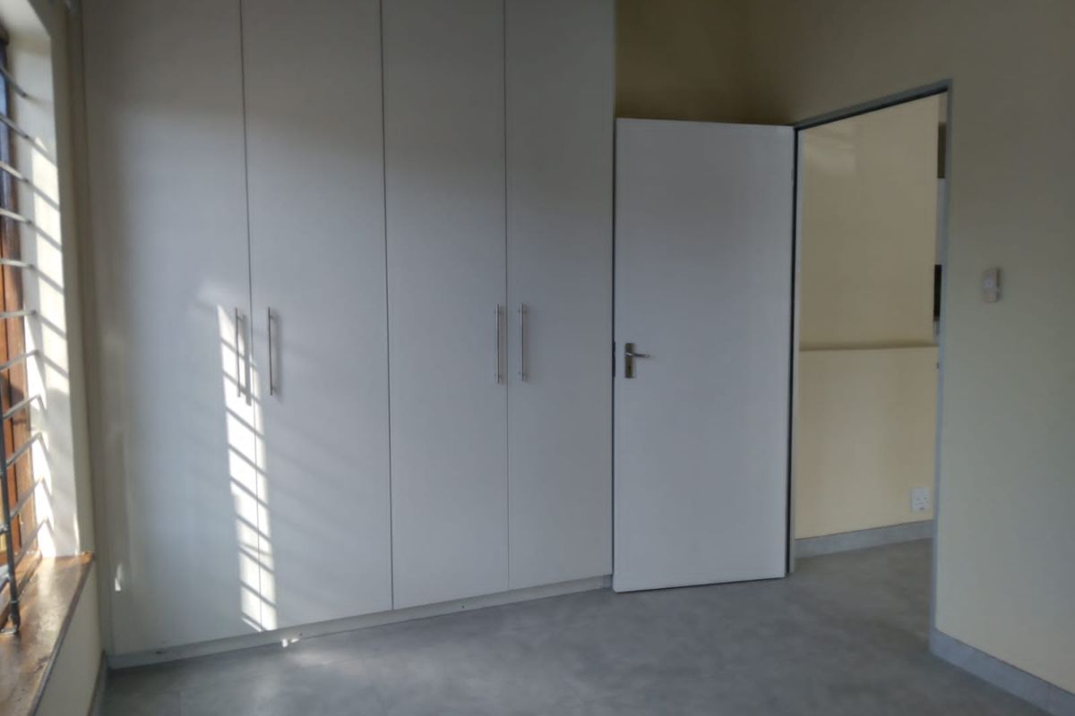 A clean, empty bedroom featuring neutral tiled flooring, built-in white cupboards, and natural light entering through a window with security bars.