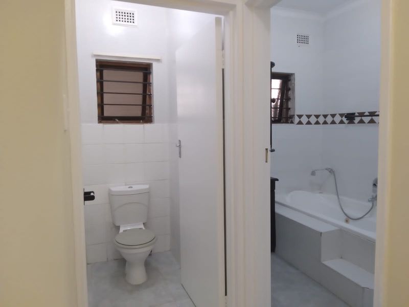 A functional bathroom and separate toilet area featuring white wall tiles, a standard bathtub with shower mixer, and ventilation grilles.