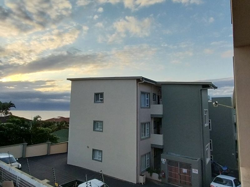 An elevated view of the apartment building exterior, featuring paved driveway areas, security fencing, and a scenic coastal horizon backdrop.