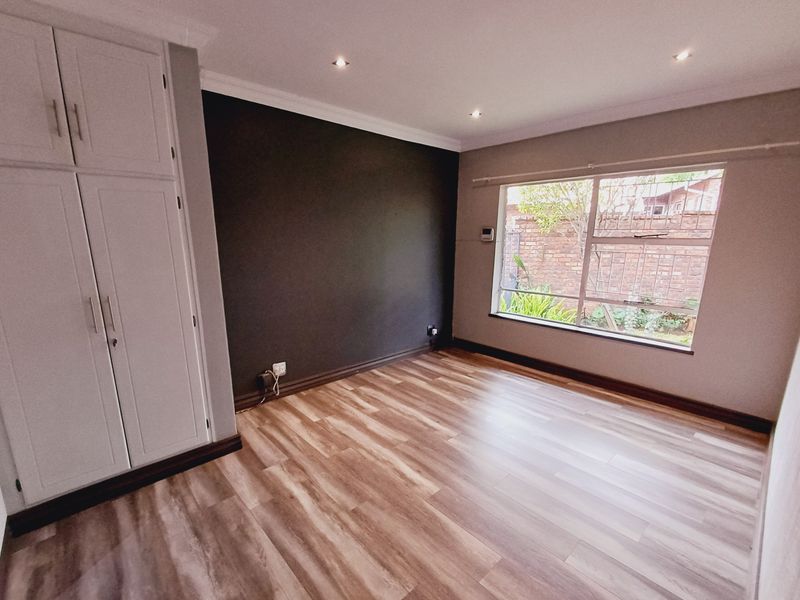 A bright, empty bedroom featuring modern laminate flooring, built-in cupboards, a dark accent wall, and recessed ceiling lighting.