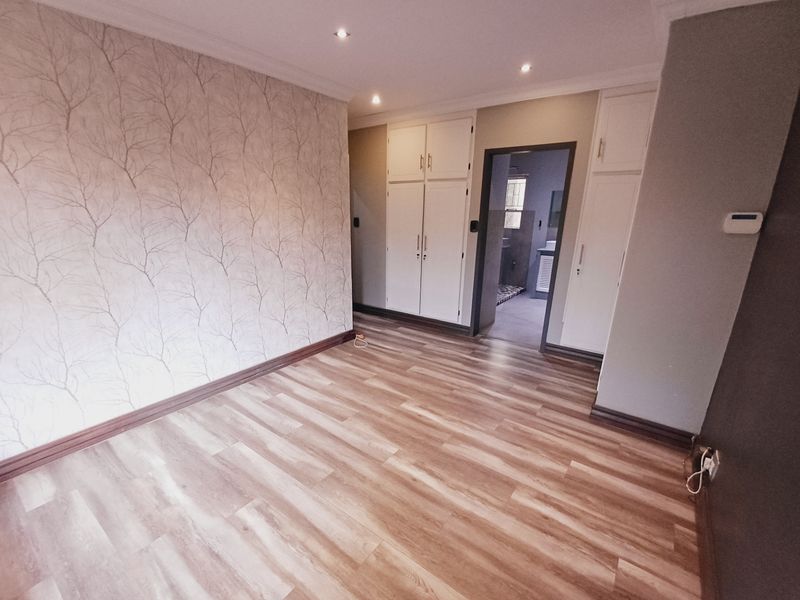 Spacious bedroom featuring modern laminate flooring, built-in cupboards, and recessed lighting, with an adjoining bathroom visible through the doorway.