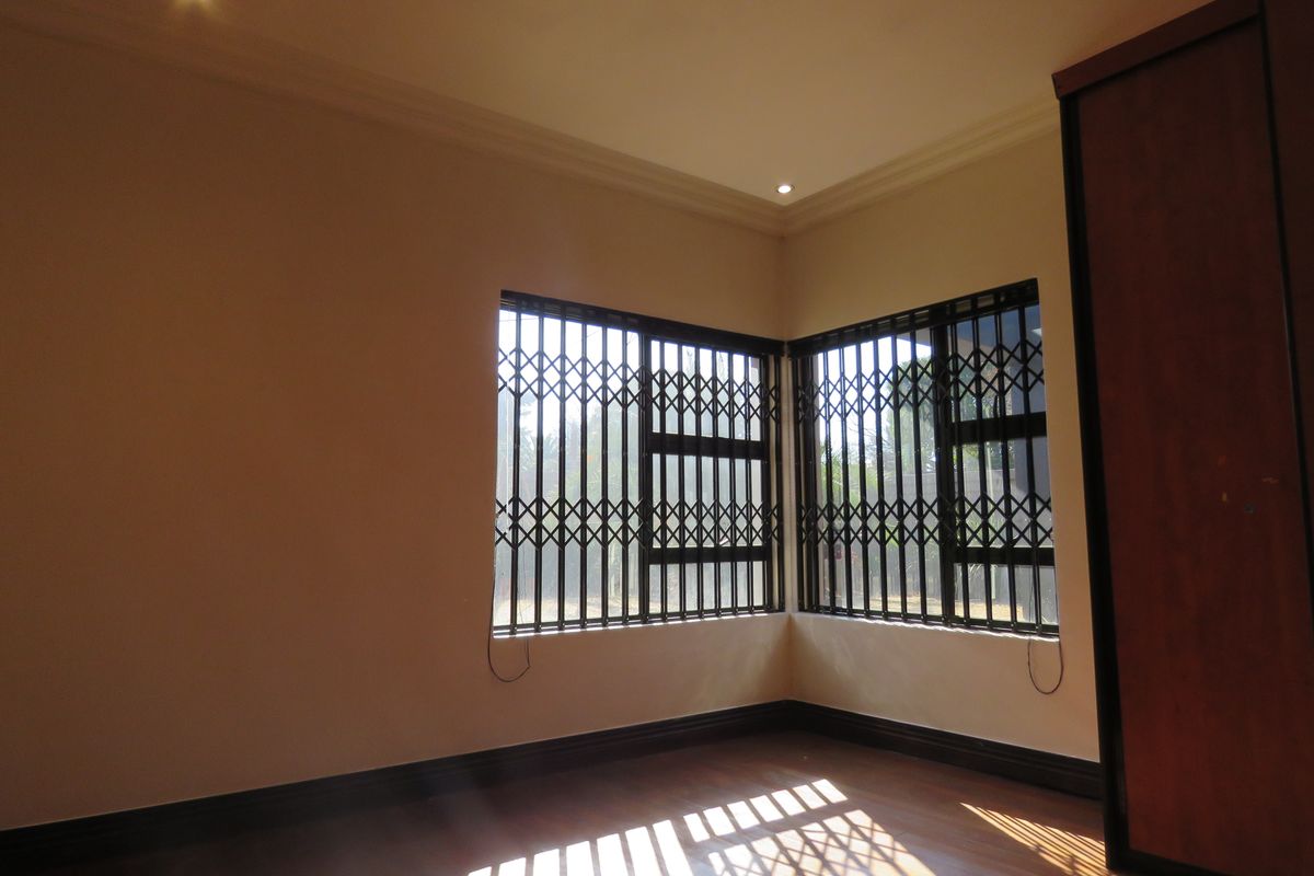 A bright, empty bedroom featuring laminate flooring, recessed downlights, and built-in wardrobes, secured with metal security gates on the corner windows.