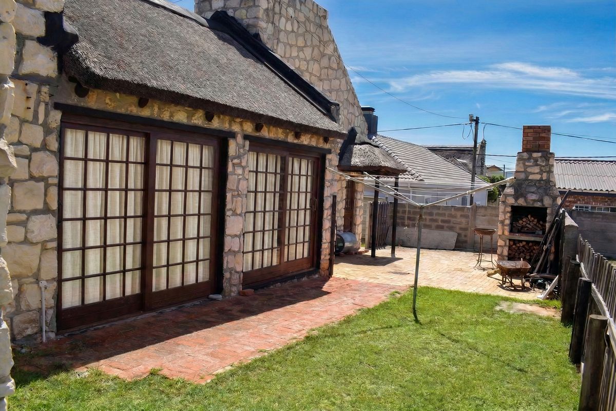 Rear exterior view featuring a thatched roof, stone-clad facade, sliding doors leading to a brick patio, and a built-in outdoor braai area.