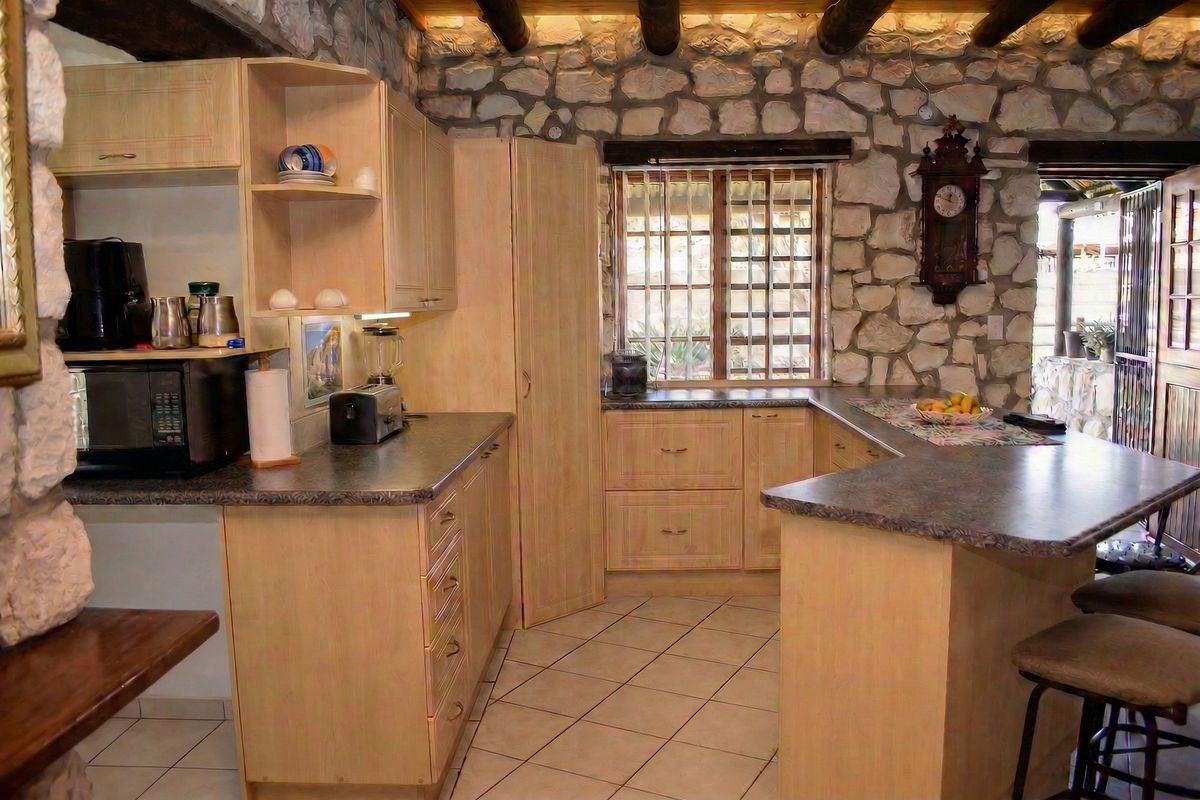 Rustic kitchen featuring stone walls, exposed timber ceiling beams, light wood cabinetry, laminate countertops, and tiled flooring.