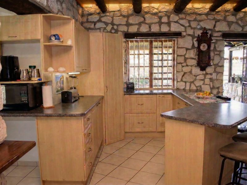 Rustic kitchen featuring stone walls, exposed timber ceiling beams, light wood cabinetry, laminate countertops, and tiled flooring.