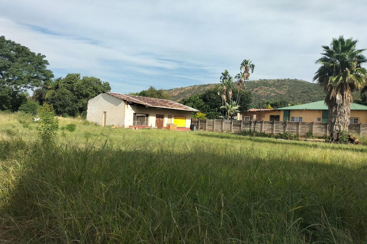 A view of the rear exterior of a residential property featuring a large overgrown yard, precast boundary wall, and a structure with a damaged roof.