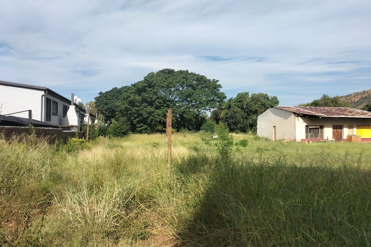 A view of a vacant residential plot featuring overgrown grass, a boundary wall, and an existing outbuilding structure in need of maintenance.