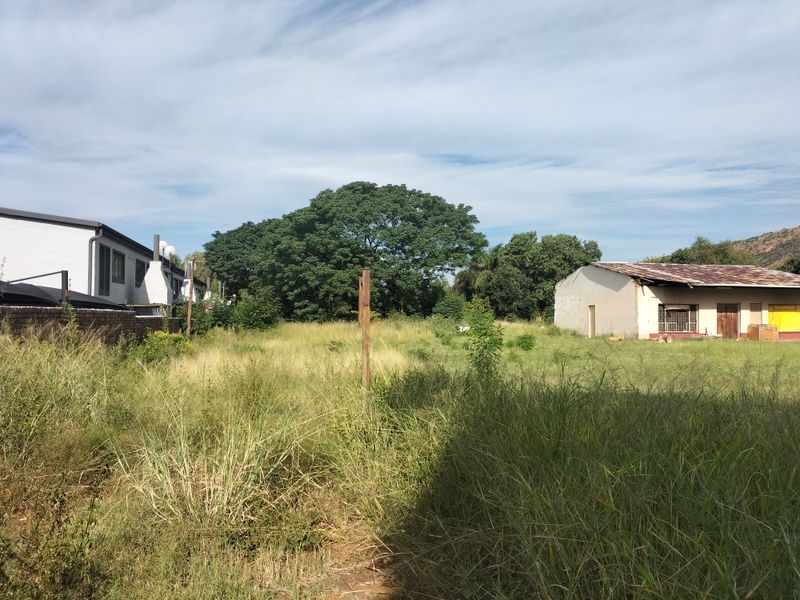 A view of a vacant residential plot featuring overgrown grass, a boundary wall, and an existing outbuilding structure in need of maintenance.