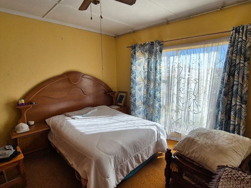 A bedroom featuring a built-in headboard, ceiling fan, and large window with curtain rails, requiring cosmetic updates to the walls and flooring.