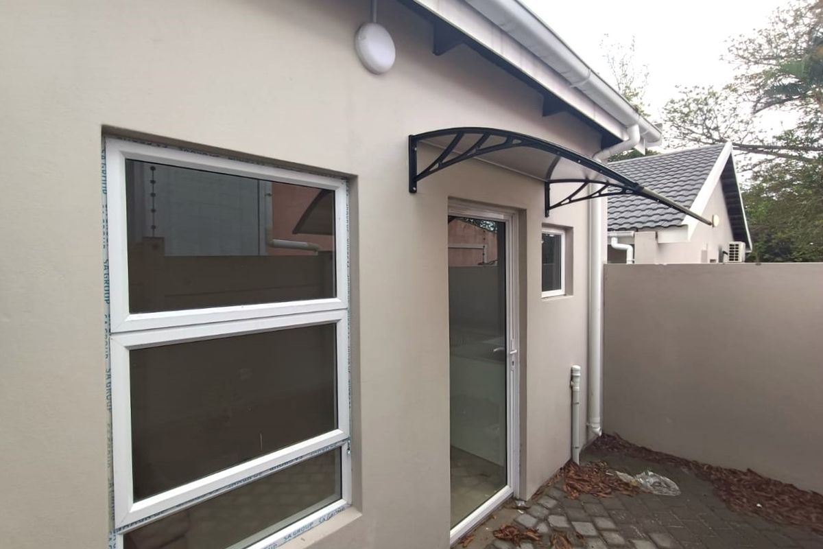 A view of the rear exterior of the flat, featuring a paved courtyard, modern aluminium window frames, a glass door, and a protective awning.